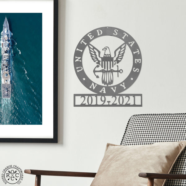 Round metal sign saying United States Navy around the edge and Navy symbol in the center with years of service etched below, hanging on wall next to photo of navy ship.