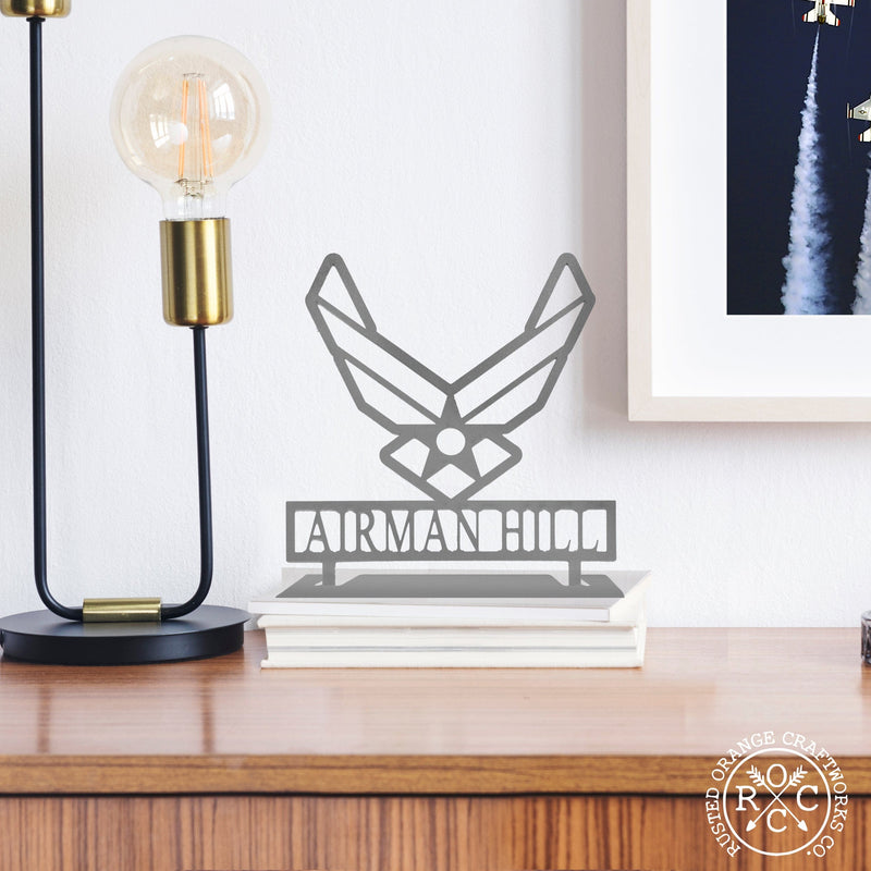 Metal US Air Force symbol with Airman Hill etched below it, sitting on desk next to lamp.