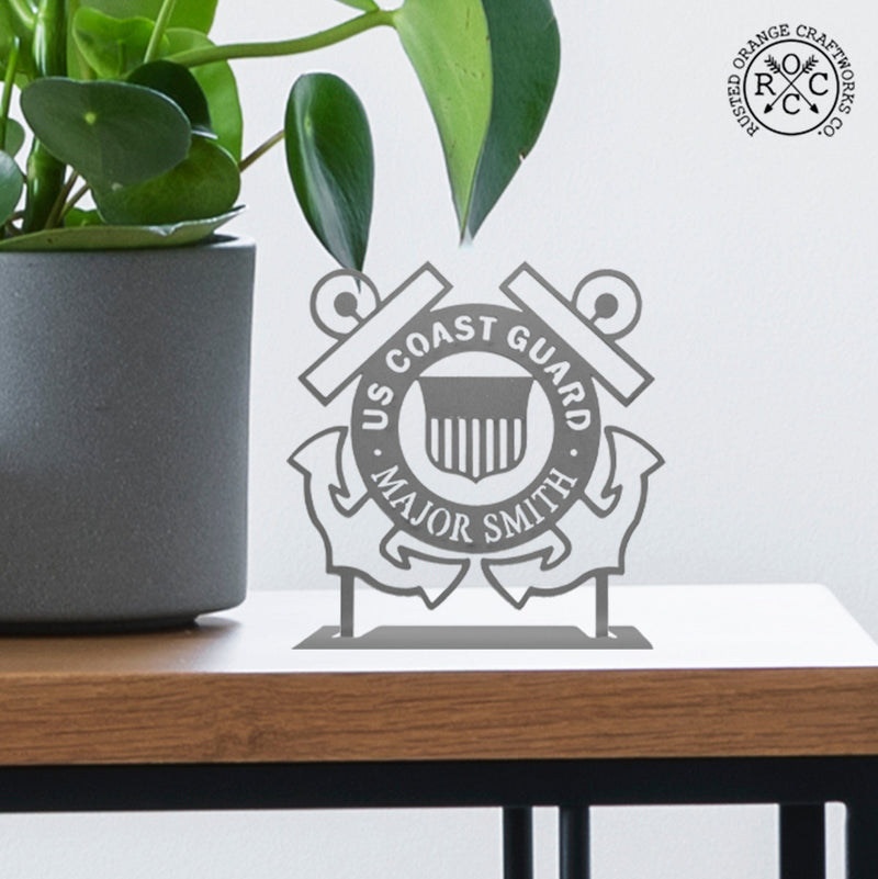Round metal sign saying US Coast Guard along top and Major Smith on bottom with 2 anchors behind and coast guard symbol in the center, sitting on shelf next to plant.