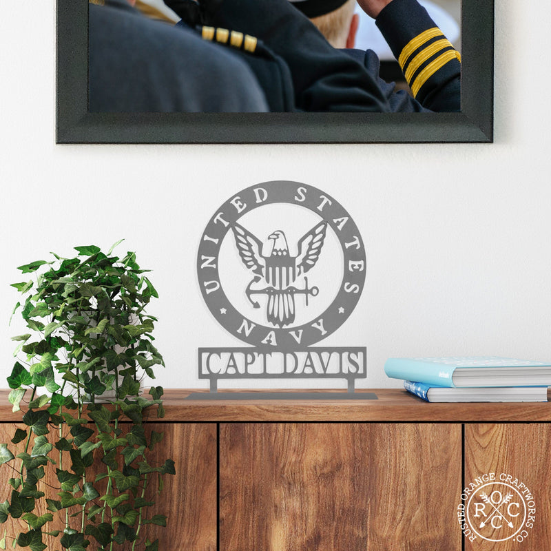 Round metal sign saying United States Navy around the edge and Navy symbol in the center with Capt Davis etched below, standing on shelf next to books and plant.