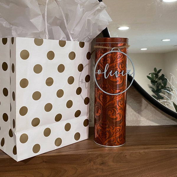 Circle metal Christmas ornament with name in center, hanging around canister next to gift bag.