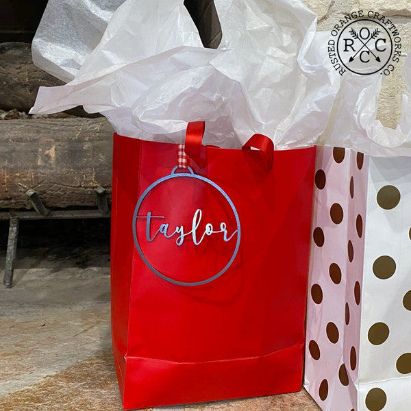 Circle metal ornament with name in center hanging on gift bag.