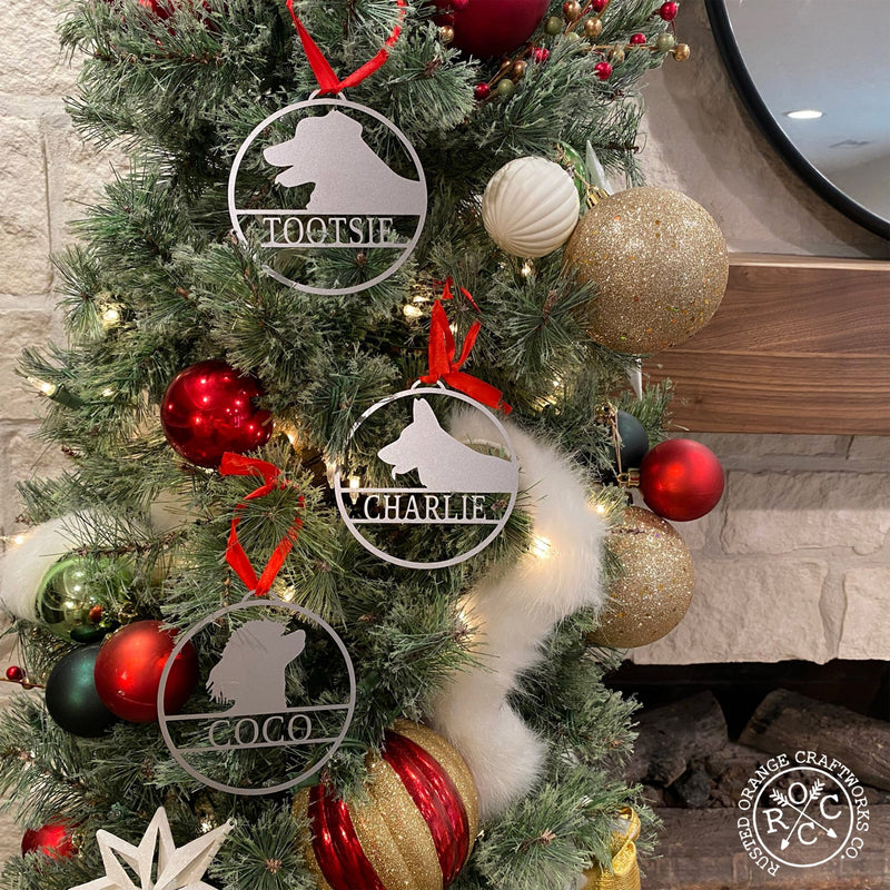 Round metal ornaments with personalized dog name and breed silhouette, hanging on Christmas tree.