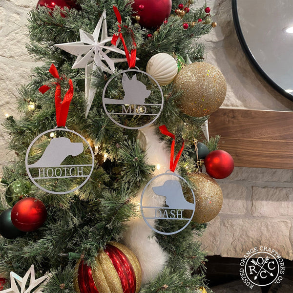 Round metal ornaments with personalized dog name and breed silhouette, hanging on Christmas tree.