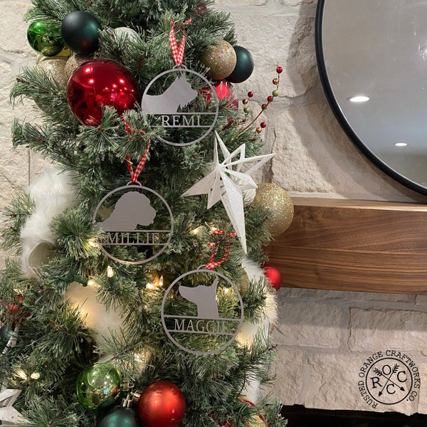 Round metal ornaments with personalized dog name and breed silhouette, hanging on Christmas tree.