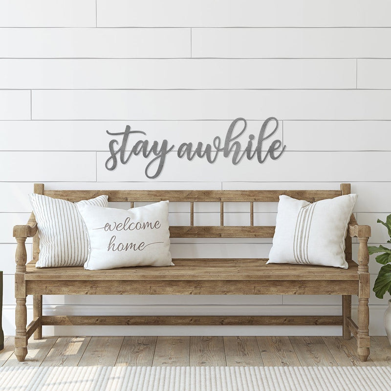 Metal sign saying stay awhile hanging outside house above bench.