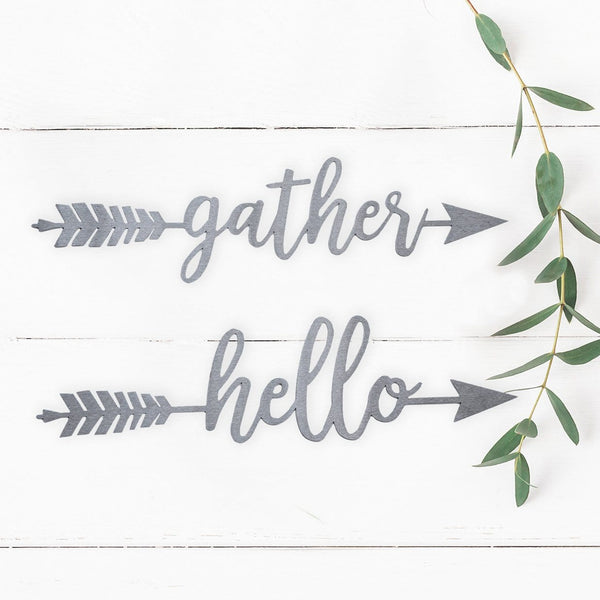 2 metal arrows with words gather and hello etched in center, hanging outside near plant.