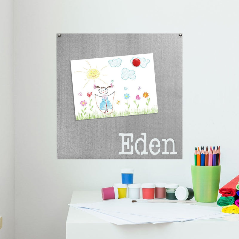 Square metal magnet board with the name Eden etched at the bottom with child's drawing held on with magnet, hanging on the wall.