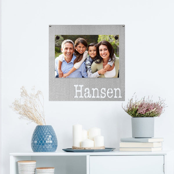 Square metal magnet board with the name Hansen etched at the bottom and family photo stuck on it, hanging on the wall above table.