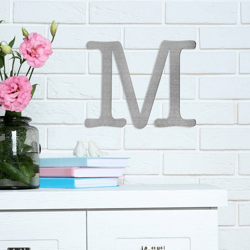 Letter M metal decoration hanging on wall above shelf with pink flowers.