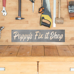 Rectangle metal sign saying Poppy's Fix it Shop hanging on wooden wall with various tools.