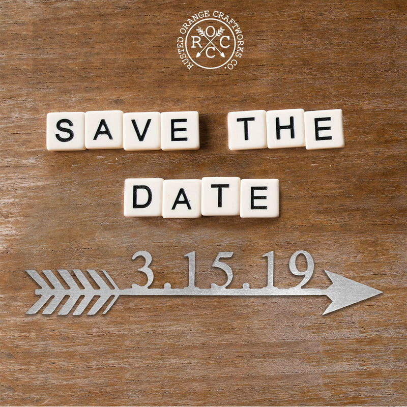 White tiles spelling out Save the Date above metal arrow with 3-15-19 on top, against wood background.