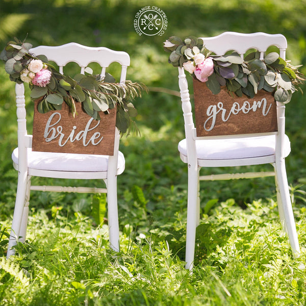 2 chairs decorated with flowers and metal signs hanging from back, one saying bride, one saying groom.