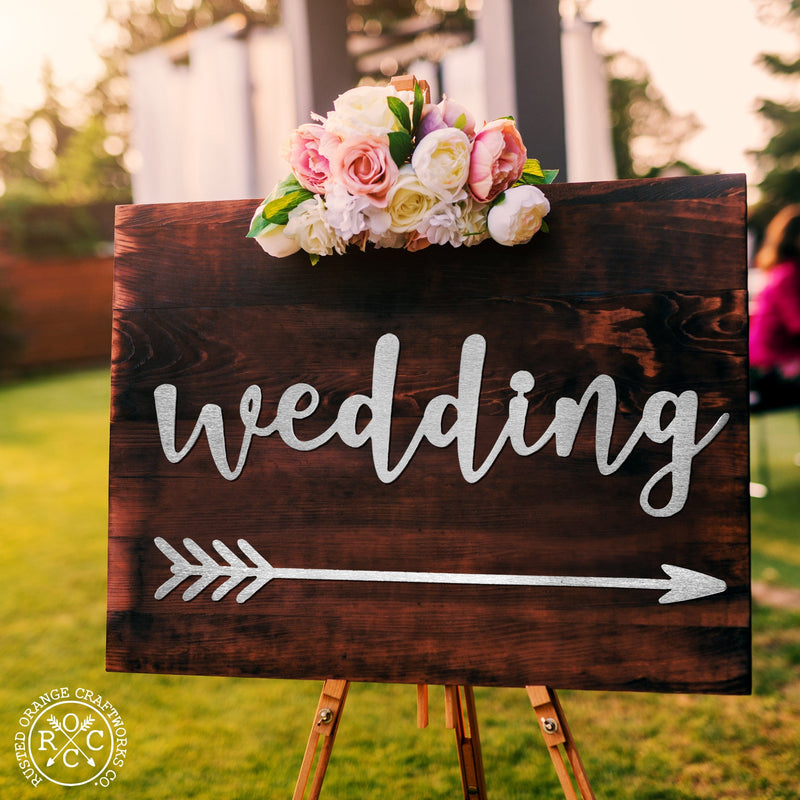 Wood base standing on easel, metal wedding sign and right-pointing arrow hanging on base under floral arrangement.