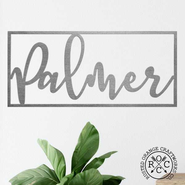 Metal cursive family name sign within rectangle hanging on wall.