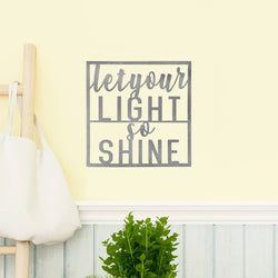 Square metal sign saying let your light so shine hanging on wall above plant.